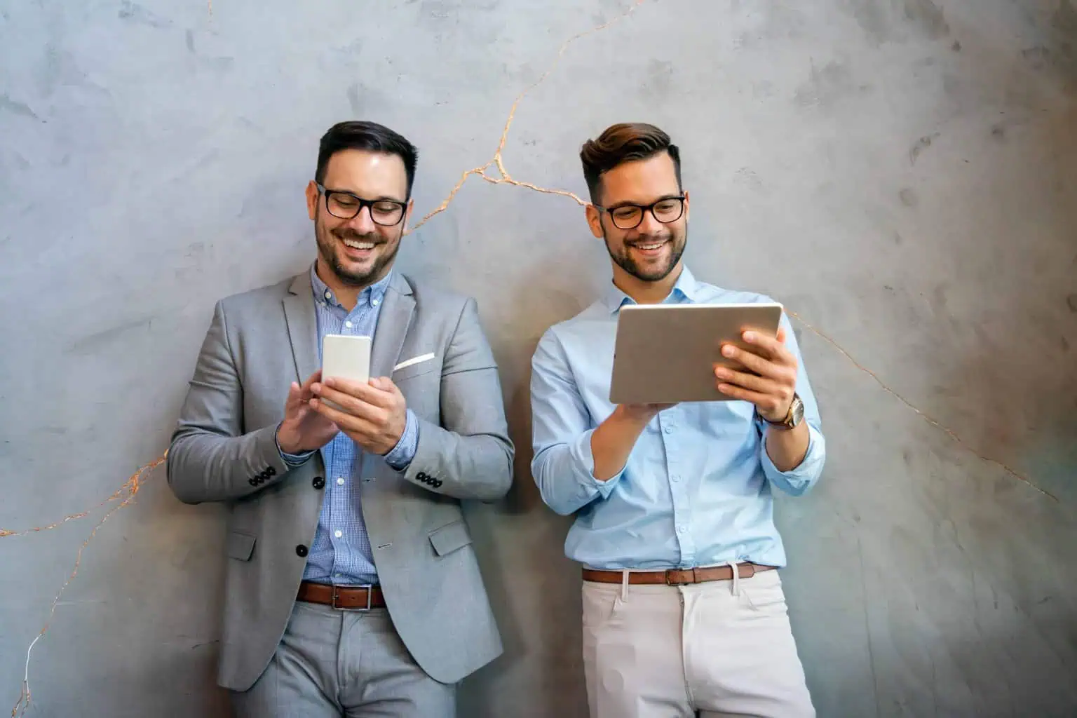 Zwei Männer in Geschäftskleidung lächeln, während sie vor einer strukturierten grauen Wand auf einem Smartphone und einem Tablet eine Immobilien-Website durchsuchen.